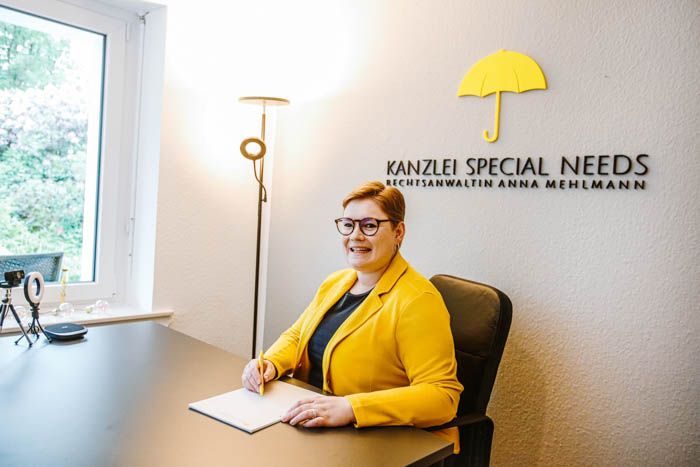 Rechtsanwältin Anna Mehlmann sitzt freundlich lächelnd an ihrem Schreibtisch in der Kanzlei. Das sonnengelbe Regenschirm-Logo der Kanzlei Special Needs prangt im Großformat an der grau gehaltenen Wand hinter ihr.