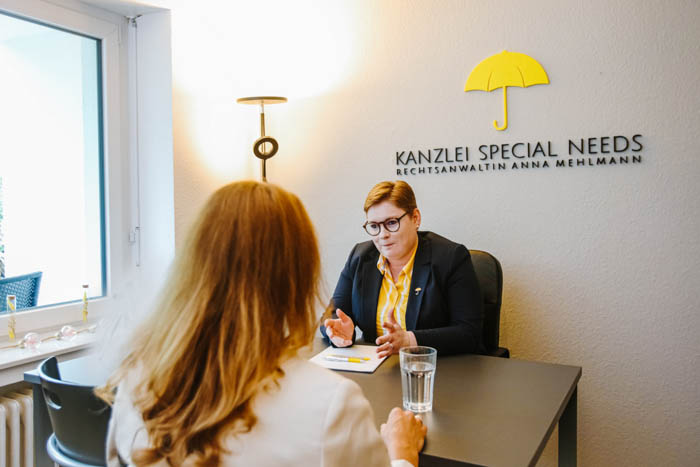 Rechtsanwältin Anna Mehlmann sitzt mit einer Mandantin in ein Gespräch vertieft am Schreibtisch. Im Hintergrund ist das gelbe Regenschirm-Logo der Kanzlei Special Needs auf grauem Hintergrund zu erkennen.